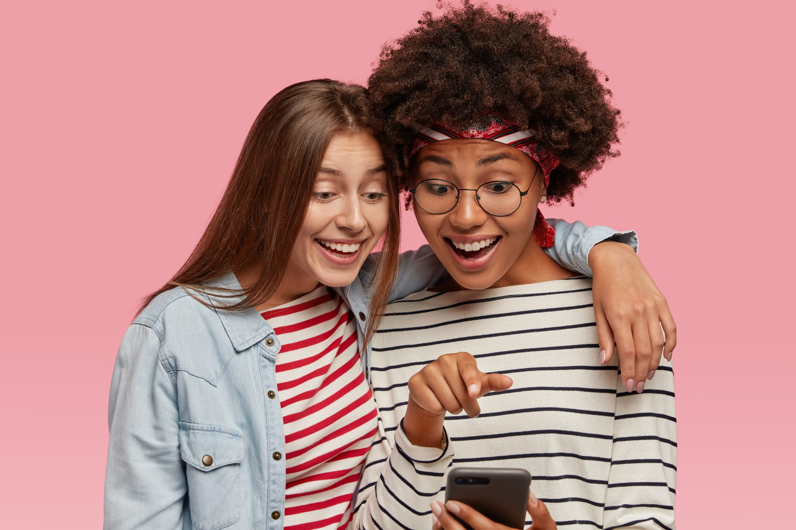 Friendly female friends have fun together, embrace and watch funny video on cellular, point at screen, dressed in striped sweaters, connected to wireless internet, isolated over pink background
